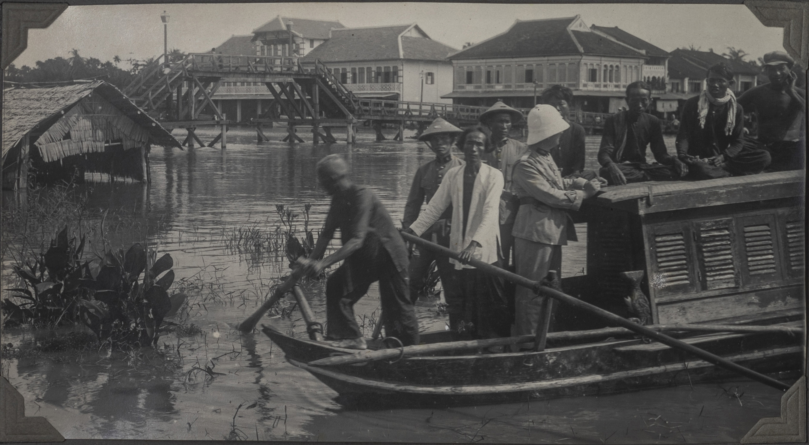 Un groupe d'hommes vietnamiens sur une pirogue arrive sur une rive. Un colon portant un casque blanc semble mener l'équipage. 